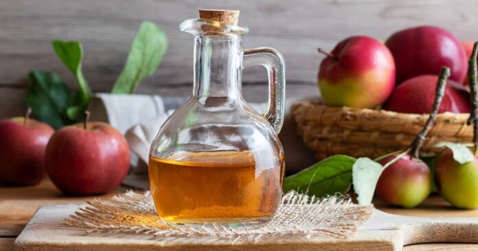 Glass bottle of apple cider vinegar in front of basket with apples