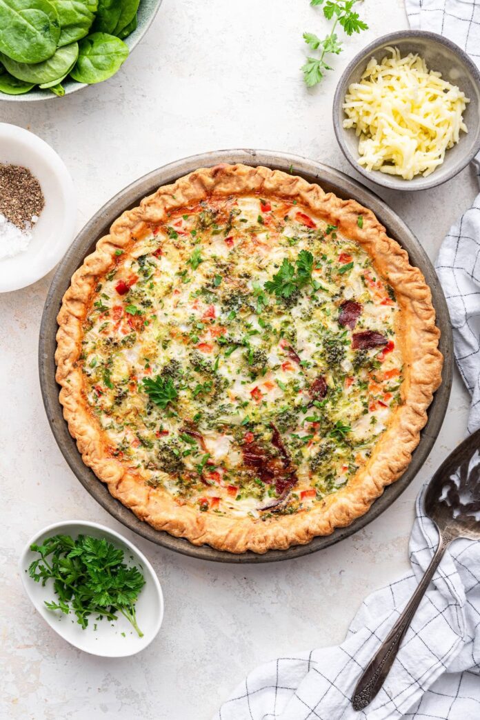 A quiche in a metal pie dish. The quiche has a golden-brown, flaky crust and is filled with a creamy mixture of egg whites, cottage cheese, and finely chopped vegetables, including broccoli, red pepper, and white onion. It also contains shredded mozzarella cheese, chopped cooked turkey bacon, and fresh herbs like parsley.