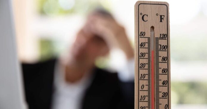 Close-up of a thermometer that reads eighty degrees. A person is in the background pressing their hand to their forehead.