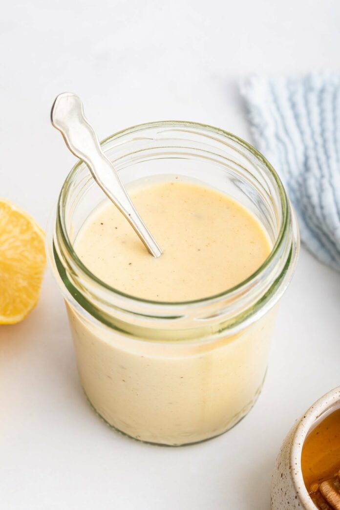 A creamy honey mustard dressing in a glass jar with a metal spoon inside the jar.