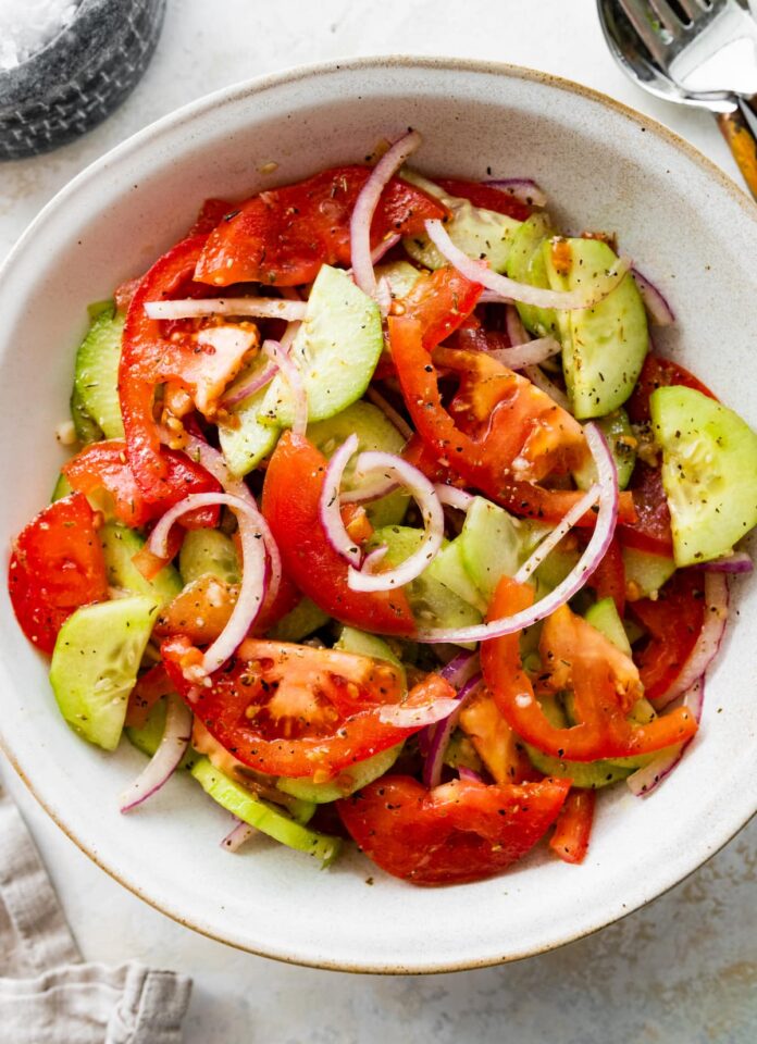 In a small white bowl, a cucumber tomato salad with sliced red onion, salt, and pepper.
