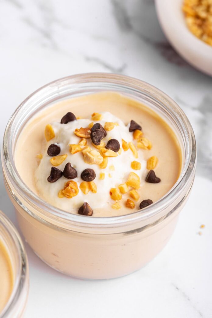 Peanut butter protein pudding in a glass container topped with mini chocolate chips, chopped peanuts, and yogurt.