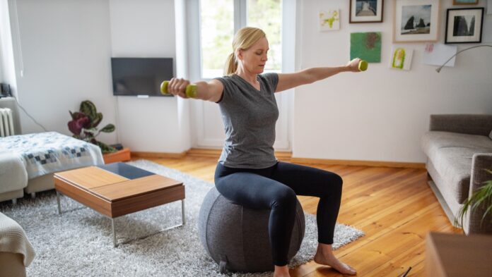 woman sitting down on a gray stability ball doing a dumbbell workout in living room